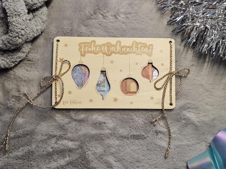 K&ouml;nnte beinhalten: Weihnachtskarte aus Holz mit ausgeschnittenen Ornamentformen, die Geldscheine zeigen. Aufschrift "Frohe Weihnachten" und "f&uuml;r Katrin". Goldene Schnur und Schleifen zur Dekoration. Die Karte ist ca. 15 cm lang.