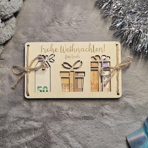 K&ouml;nnte beinhalten: Eine Weihnachtsgeschenkkarte aus Holz mit ausgeschnittenen Geschenkformen und dem deutschen Text "Frohe Weihnachten!" und "fur Leah". Die Karte enth&auml;lt Euro-Banknoten und ist mit Schnurschleifen gebunden. Der Hintergrund ist eine graue, verschwommene Oberfl&auml;che.