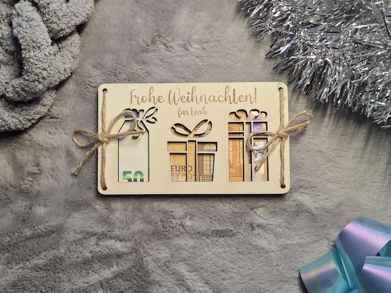 K&ouml;nnte beinhalten: Eine Weihnachtsgeschenkkarte aus Holz mit ausgeschnittenen Geschenkformen und dem deutschen Text "Frohe Weihnachten!" und "fur Leah". Die Karte enth&auml;lt Euro-Banknoten und ist mit Schnurschleifen gebunden. Der Hintergrund ist eine graue, verschwommene Oberfl&auml;che.