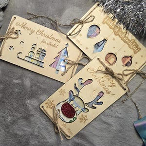 K&ouml;nnte beinhalten: Weihnachtsgeschenkanh&auml;nger aus Holz mit ausgeschnittenen Designs und Bindf&auml;den. Ein Anh&auml;nger sagt "Merry Christmas" und "f&uuml;r Isabel", ein anderer sagt "Frohe Weihnachten!", und ein dritter zeigt ein Rentier-Design. Die Anh&auml;nger haben ausgeschnittene Formen von Weihnachtsschmuck und einem Weihnachtsbaum.