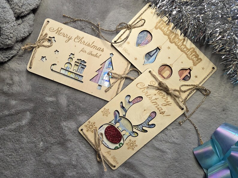 K&ouml;nnte beinhalten: Weihnachtsgeschenkanh&auml;nger aus Holz mit ausgeschnittenen Designs und Bindf&auml;den. Ein Anh&auml;nger sagt "Merry Christmas" und "f&uuml;r Isabel", ein anderer sagt "Frohe Weihnachten!", und ein dritter zeigt ein Rentier-Design. Die Anh&auml;nger haben ausgeschnittene Formen von Weihnachtsschmuck und einem Weihnachtsbaum.