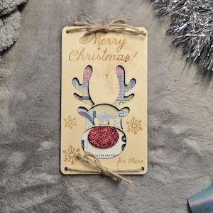 K&ouml;nnte beinhalten: Eine Weihnachtsgeschenkkarte aus Holz mit einem Rentier-Design. Die Karte tr&auml;gt die Aufschrift "Merry Christmas!" und hat eine rote Glitzernase. Das Geweih des Rentiers ist ausgeschnitten und gibt das Geld im Inneren frei.