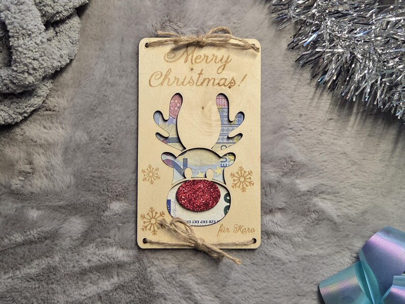 K&ouml;nnte beinhalten: Eine Weihnachtsgeschenkkarte aus Holz mit einem Rentier-Design. Die Karte tr&auml;gt die Aufschrift "Merry Christmas!" und hat eine rote Glitzernase. Das Geweih des Rentiers ist ausgeschnitten und gibt das Geld im Inneren frei.