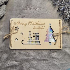 K&ouml;nnte beinhalten: Weihnachtskarte aus Holz mit dem Text "Merry Christmas f&uuml;r Isabel". Die Karte zeigt ausgeschnittene Designs eines Weihnachtsbaums, Geschenke und Sterne, die mit Bindfaden gebunden sind.