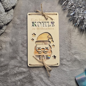 K&ouml;nnte beinhalten: Ein Holz-Geschenkanh&auml;nger mit einem Weihnachtsmann-Design, mit ausgeschnittenen Sternen und einem Gesicht. Der Anh&auml;nger tr&auml;gt den deutschen Text "Ein bisschen K&Ouml;LLE zu Weihnachten" und ist mit Bindfaden gebunden. Das Gesicht des Weihnachtsmanns ist mit Geld gef&uuml;llt.