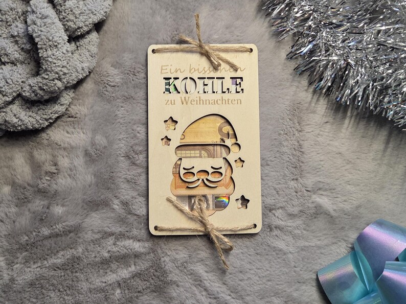 K&ouml;nnte beinhalten: Ein Holz-Geschenkanh&auml;nger mit einem Weihnachtsmann-Design, mit ausgeschnittenen Sternen und einem Gesicht. Der Anh&auml;nger tr&auml;gt den deutschen Text "Ein bisschen K&Ouml;LLE zu Weihnachten" und ist mit Bindfaden gebunden. Das Gesicht des Weihnachtsmanns ist mit Geld gef&uuml;llt.