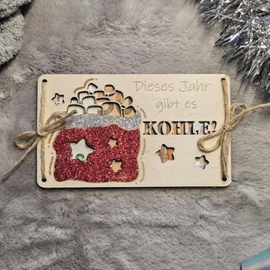 K&ouml;nnte beinhalten: Weihnachtsbaumschmuck aus Holz mit dem deutschen Text "Dieses Jahr gibt es KOHLE?" mit einem rot glitzernden Sack mit Sternaussparungen. Der Schmuck ist mit Bindfaden gebunden.