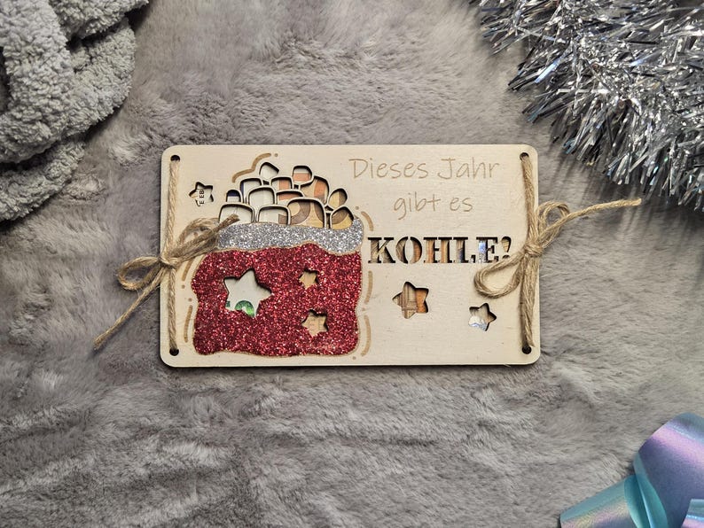 K&ouml;nnte beinhalten: Weihnachtsbaumschmuck aus Holz mit dem deutschen Text "Dieses Jahr gibt es KOHLE?" mit einem rot glitzernden Sack mit Sternaussparungen. Der Schmuck ist mit Bindfaden gebunden.