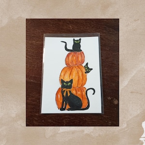 May include: A Halloween-themed illustration featuring three black cats perched on a stack of three orange pumpkins. The cats have green eyes and are in various poses. One cat wears a yellow bow tie. The artwork is on a white background.