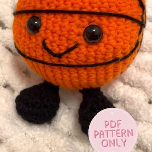 May include: A crocheted basketball with black accents, black eyes, and small black feet. The orange ball has black lines and a smiling face. The text "Basketball pattern" is at the top. A pink circle with the text "PDF PATTERN ONLY" is at the bottom.