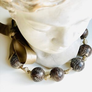 May include: A brown wooden bead necklace with a gold ribbon tied around the beads. The necklace is displayed on a white mannequin bust.