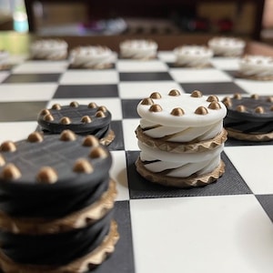 May include: Close-up of a checkerboard with black and white squares. Stacks of checkers are visible, alternating between black and white. The checkers have gold-colored trim and small gold studs on top, creating a classic board game aesthetic.