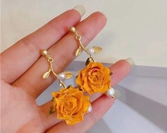 May include: A pair of gold-colored earrings featuring bright yellow rose pendants. Each earring has a small pearl and gold-colored leaves. The earrings are held up by a gold-colored hook.