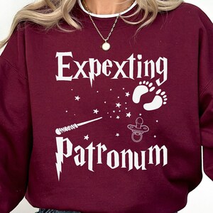 May include: A maroon sweatshirt with white text that reads "Expecting Patronum" with a wand, stars, footprints, and a pacifier graphic.