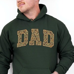 May include: Forest green hoodie with the word "DAD" in a camouflage pattern across the chest. The hoodie has a front pocket and a drawstring hood. The sleeves are long, and the material appears to be a soft fabric.