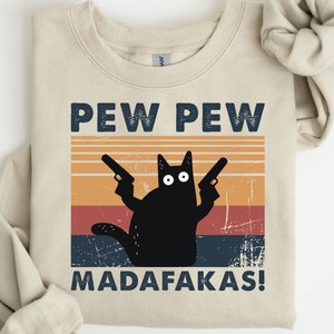 May include: A beige sweatshirt with a black cat holding two guns and the text "PEW PEW MADAFAKAS!" in a vintage style graphic.