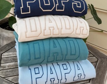 May include: A stack of four folded sweatshirts in navy, cream, teal, and light blue. Each sweatshirt features a different embroidered word: "PUPS", "DAD", "DAD", and "PAPA" in a collegiate style font. The sweatshirts are stacked on a wooden surface.