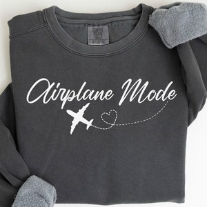 May include: Charcoal grey sweatshirt featuring the text "Airplane Mode" in white script, a white aeroplane graphic, and a dotted heart trail. The sleeves are cuffed. A "Comfort Colors" label is in the bottom right corner.