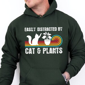 May include: A dark green Gildan hoodie with the text "Easily Distracted By Cat & Plants" in white. The text is surrounded by a rainbow-colored stripe design. A white silhouette of a cat is shown reaching for a plant in a pot.