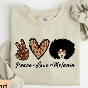 May include: A beige sweatshirt with a peace sign, heart, and a woman with an afro wearing heart-shaped sunglasses. The text "Peace Love Melanin" is printed below the images.