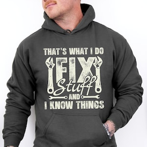 May include: Dark heather grey hoodie with a white graphic that says "That's what I do I fix stuff and I know things" with wrench illustrations.
