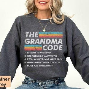 May include: Heather grey sweatshirt with the text "THE GRANDMA CODE" in a vintage style font. The text is surrounded by a colorful stripe design. Below the text is a list of five rules: "1. BEDTIME IS WHENEVER", "2. THE ANSWER IS ALWAYS YES", "3. I WILL ALWAYS HAVE YOUR BACK", "4. MOM DOESN'T NEED TO KNOW", "5. HUGS ARE MANDATORY".