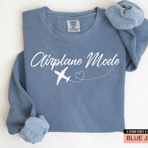 May include: A blue jean-coloured sweatshirt featuring the words "Airplane Mode" in white script, a white airplane graphic, and a dotted heart trail. The sweatshirt has a crew neck and long sleeves.