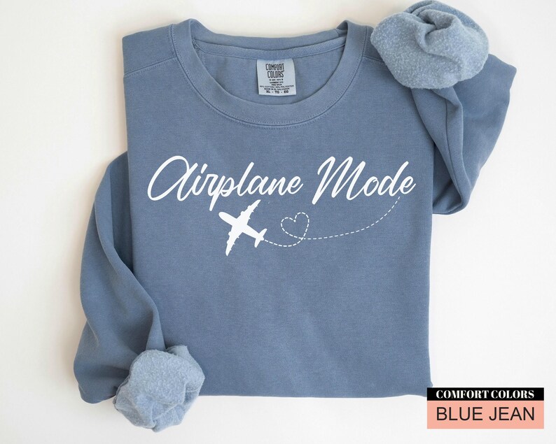 May include: A blue jean-coloured sweatshirt featuring the words "Airplane Mode" in white script, a white airplane graphic, and a dotted heart trail. The sweatshirt has a crew neck and long sleeves.