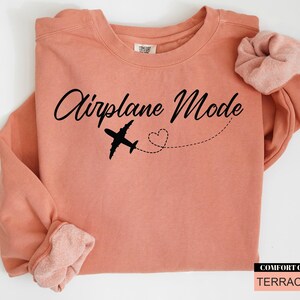 May include: A terracotta-coloured sweatshirt with the words "Airplane Mode" in a cursive font. A black airplane graphic and a dotted line with a heart shape are below the text. A matching scrunchie rests on the shoulder.