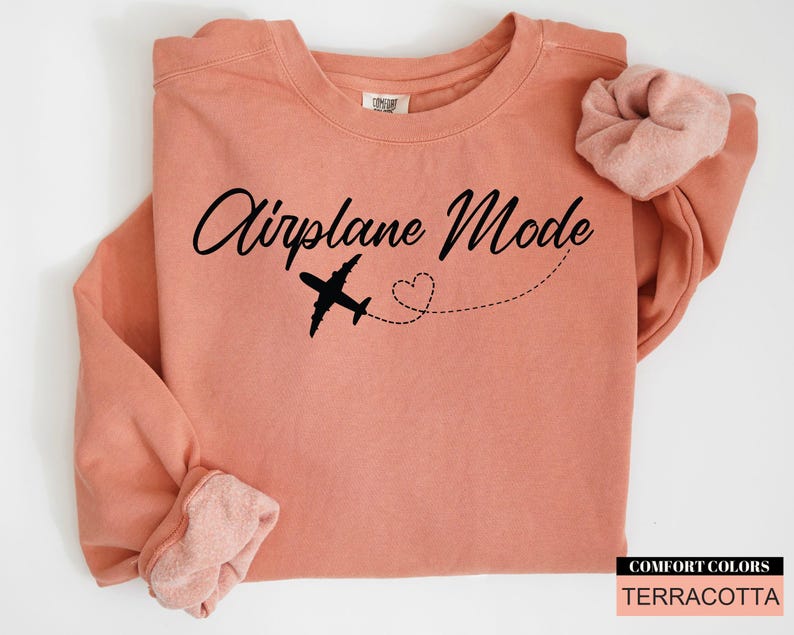 May include: A terracotta-coloured sweatshirt with the words "Airplane Mode" in a cursive font. A black airplane graphic and a dotted line with a heart shape are below the text. A matching scrunchie rests on the shoulder.
