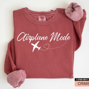 May include: Crimson-coloured sweatshirt featuring the text "Airplane Mode" in white script, a white aeroplane graphic, and a dotted heart. The sweatshirt has rolled-up sleeves and a Comfort Colors label.