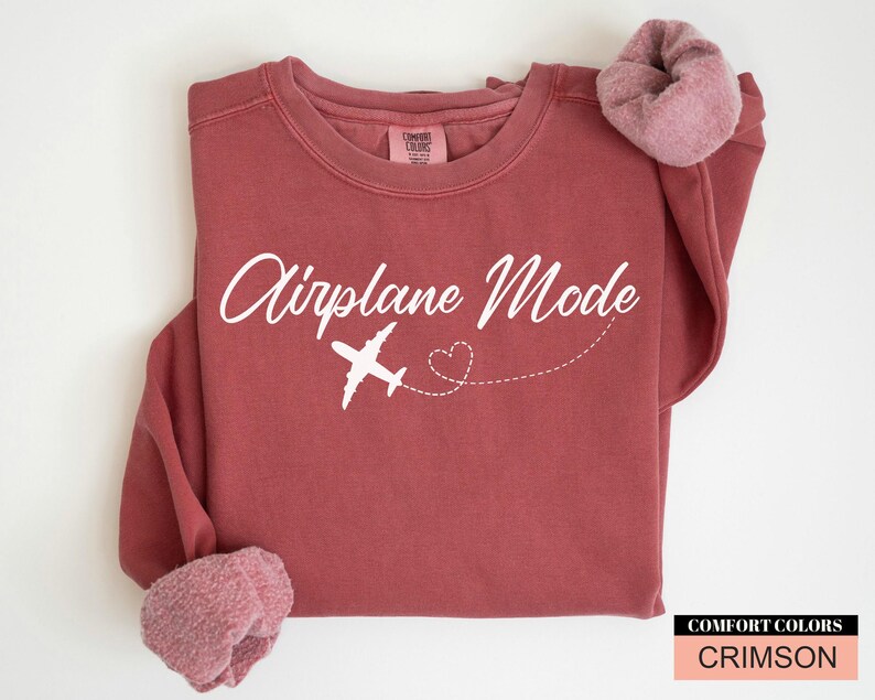May include: Crimson-coloured sweatshirt featuring the text "Airplane Mode" in white script, a white aeroplane graphic, and a dotted heart. The sweatshirt has rolled-up sleeves and a Comfort Colors label.