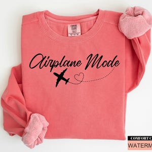 May include: A coral-coloured sweatshirt featuring the text "Airplane Mode" in a cursive script, a black aeroplane graphic, and a dotted heart trail. The sleeves are cuffed. The Comfort Colors brand and colour name are visible.