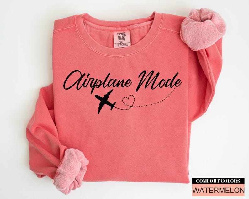 May include: A coral-coloured sweatshirt featuring the text "Airplane Mode" in a cursive script, a black aeroplane graphic, and a dotted heart trail. The sleeves are cuffed. The Comfort Colors brand and colour name are visible.