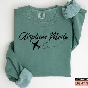 May include: Light green sweatshirt featuring the text "Airplane Mode" in black script, a black airplane graphic, and a dotted heart trail. The sweatshirt is folded, with the sleeves visible. The Comfort Colors brand is visible.