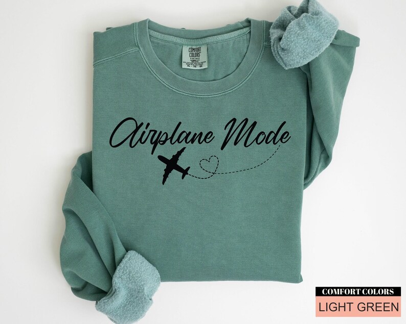 May include: Light green sweatshirt featuring the text "Airplane Mode" in black script, a black airplane graphic, and a dotted heart trail. The sweatshirt is folded, with the sleeves visible. The Comfort Colors brand is visible.