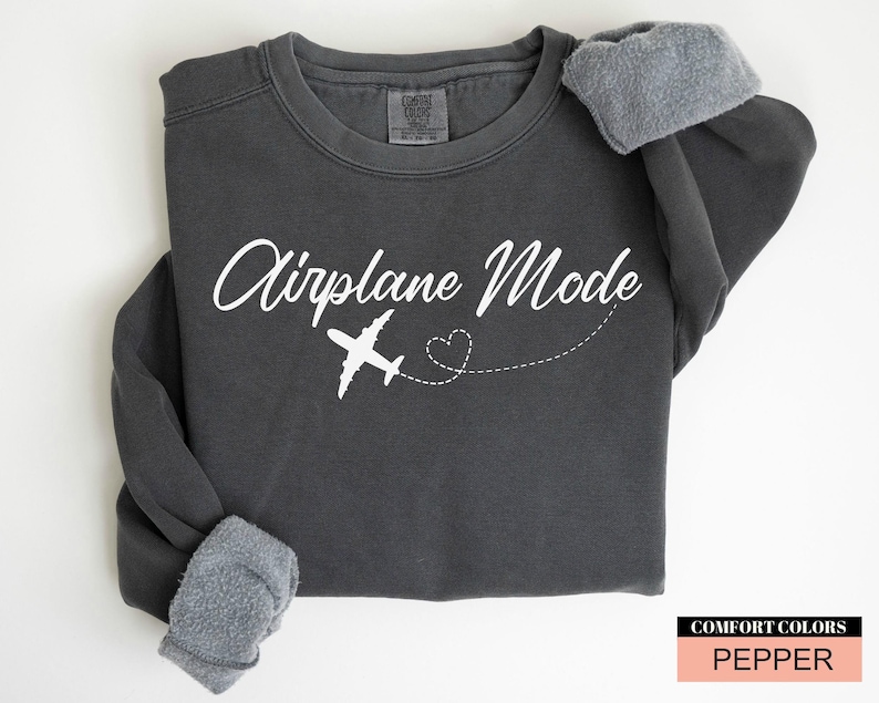May include: Charcoal grey sweatshirt featuring the text "Airplane Mode" in white script, a white aeroplane graphic, and a dotted heart trail. The sleeves are cuffed. A "Comfort Colors" label is in the bottom right corner.