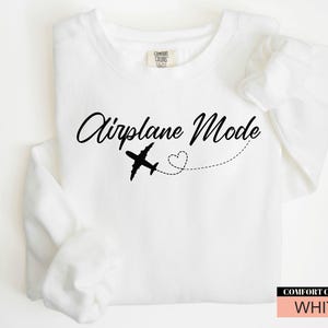 May include: White sweatshirt featuring the text "Airplane Mode" in a cursive script, a black airplane illustration, and a dotted line with a heart. The sweatshirt is labelled "Comfort Colors" and is white.