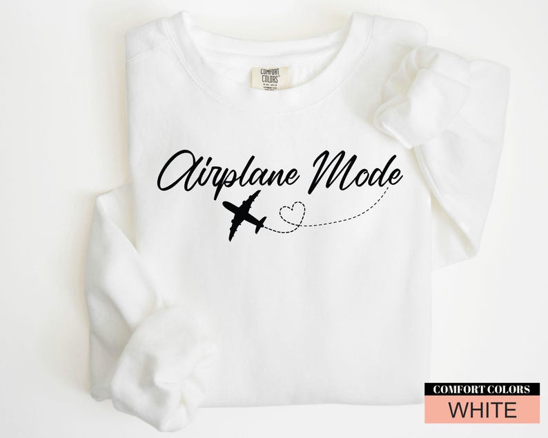 May include: White sweatshirt featuring the text "Airplane Mode" in a cursive script, a black airplane illustration, and a dotted line with a heart. The sweatshirt is labelled "Comfort Colors" and is white.
