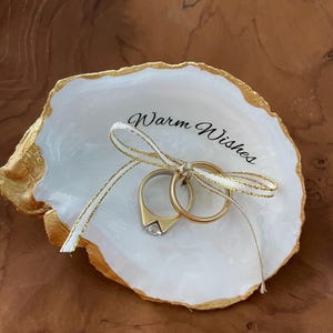 May include: White ceramic ring dish with a gold rim and the words "Warm Wishes" inscribed. Two gold wedding bands are placed in the dish with a white and gold ribbon tied around them.