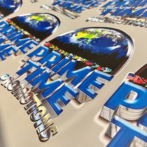 Pu&ograve; includere: Primo piano di un mucchio di adesivi blu e argento con il testo "PRIME TIME PROMOTIONS" e una grafica di un globo terrestre.