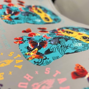 Pu&ograve; includere: Primo piano di un design stampato con un teschio turchese con farfalle colorate e motivi floreali. Il testo "Stay True" &egrave; visibile nell'angolo in basso a destra.