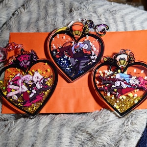 May include: Three heart-shaped keychains with glitter and cartoon characters inside. The keychains are black with gold glitter and feature a couple in each heart.
