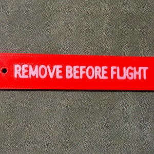 May include: A bright red rectangular tag with the words "REMOVE BEFORE FLIGHT" in white, bold letters. The tag has a small hole on the left side. The tag is likely used in aviation.
