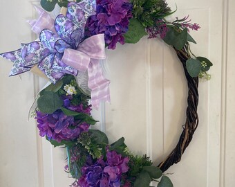 May include: A purple and white wreath featuring a large bow and hydrangea flowers. The wreath is crafted from natural materials and is ideal for spring or summer decoration.