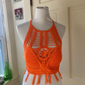 Puede incluir: Top halter de ganchillo naranja brillante con flecos. El top hecho a mano presenta un diseño floral y un cuello halter. El top se muestra en un maniquí blanco.