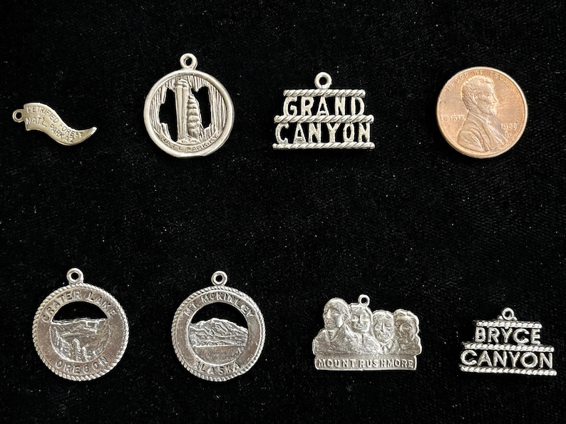 May include: A collection of silver charm pendants featuring various American landmarks, including the Grand Canyon, Mount Rushmore, Crater Lake, and Bryce Canyon. The charms are all in a circular or rectangular shape and have a silver finish.