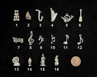 Dijes musicales de plata de ley: instrumentos musicales, notas, símbolos de clave de sol y de fa, arpa, xilófono, victrola, bajo y micrófono.