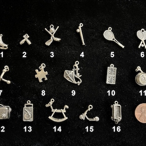 May include: A collection of silver-toned charms, including a figure skate, a pair of ballet shoes, hockey sticks, a baseball bat, a tennis racket, and a pair of tennis rackets. Other charms include jacks, a puzzle piece, a kite, dominoes, a music box, a radio, a rocking horse, a scooter, and a camera.