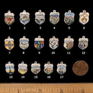 May include: A collection of 17 enamel and silver charms, each depicting a different state or location. The charms are arranged in rows of four, with a single charm at the bottom. The charms are all in the shape of a shield, and each one features a different design. The charms are all in good condition, and they would make a great addition to any charm bracelet or necklace.